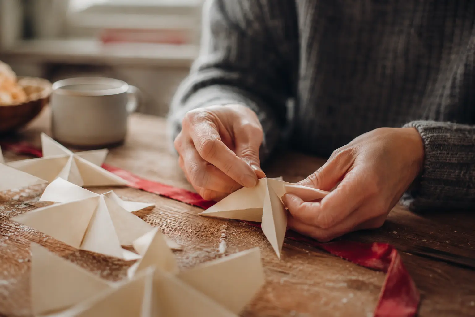 Adult hands folding simple cream paper stars at a real kitchen table with a narrow red cotton ribbon beneath them